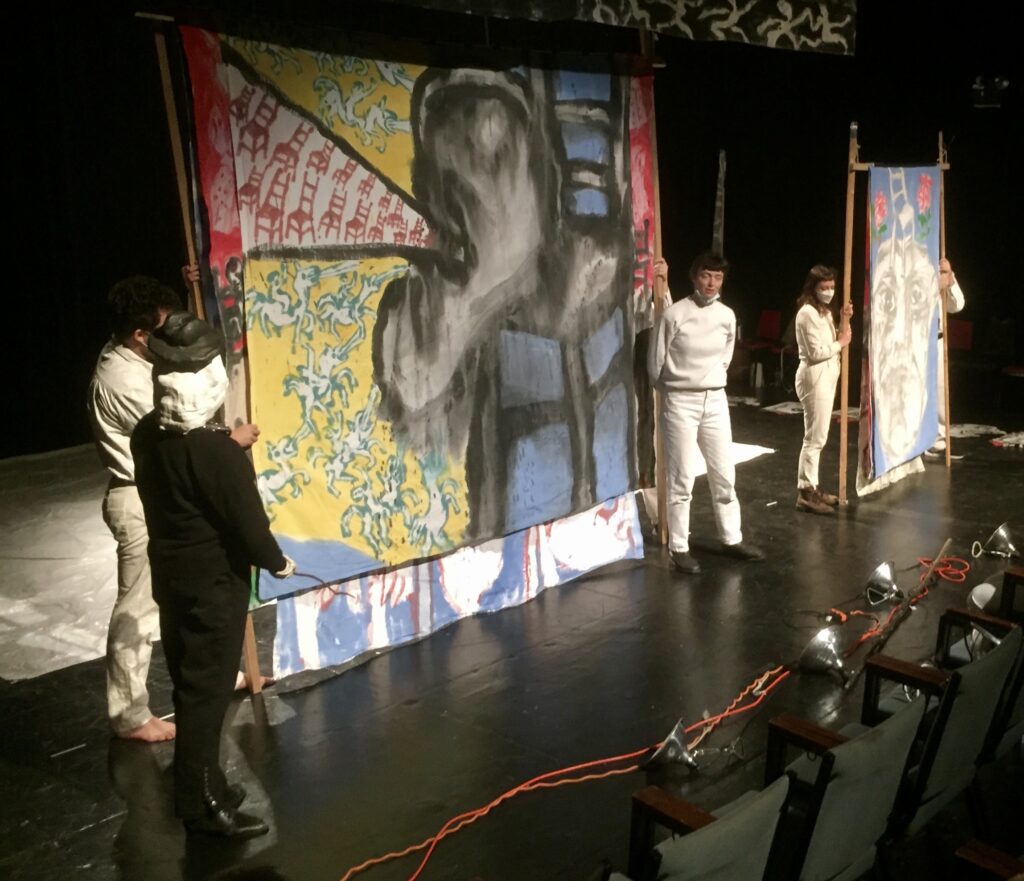 Figure 6. Bread and Puppet Theater company members (l. to r.: Joshua Krugman, Barbara Leber [in mask as Bob the Butcher], Amelia Castillo, Esteli Kitchen, and Joe Therrien) rehearsing a cantastoria version of a series of bedsheet paintings mounted on poles and crosspieces, for The Persians at Theater for the New City in New York City, December 2021. (Photo: John Bell)
